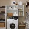 A white cabinet with three doors is shown, filled with various bathroom or laundry items. To the left, a tall shelving unit holds folded towels and baskets. A washing machine is positioned below the cabinet. Náhled obrázku produktu