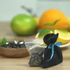 A close-up shot of a small black pouch tied with a blue ribbon, next to a clear plastic bag filled with dark, irregular-shaped stones. In the blurred background, there are fruits like an apple, an orange, and lemons, suggesting a natural or health-related  Anteprima dell’immagine prodotto