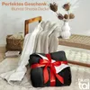 A dark gray sherpa blanket is folded and tied with a red ribbon, presented as a gift. A lighter colored sherpa blanket is draped over a chair in the background, and a woven basket with blankets is also visible. Anteprima dell’immagine prodotto