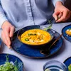 A person is garnishing a bowl of soup with a sprig of greens, with other dishes and a glass of water on the table. Voir les images du produit