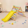 A colorful wooden climbing triangle with a yellow slide, set up on a carpeted floor in a room. Several small colorful balls are scattered around the base of the structure. Produktbild-Vorschau