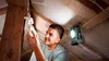 A man is changing a light bulb in an attic, illuminated by a work light. Anteprima dell’immagine prodotto