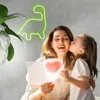 A mother is receiving a card from her daughter, who is kissing her cheek. A neon dinosaur light is visible in the background. Podgląd zdjęcia produktu