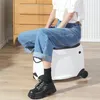 A person is sitting on a white rolling suitcase in a room with wooden furniture and a striped wall. Produktbild-Vorschau