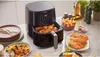 A Philips Airfryer is shown on a kitchen counter, surrounded by prepared food items like chicken wings, salmon, and vegetables. Produktbild-Vorschau