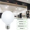 A large, white globe LED light bulb is shown in the foreground, with a kitchen or dining area in the background featuring pendant lights and cabinetry. Produktbild-Vorschau