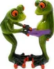 Two green frog figurines with large orange eyes are standing and holding a purple object between them. Produktbild-Vorschau