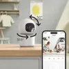 A white security camera is shown on a wooden table, with a smartphone displaying its feed next to it. The background shows a nursery or child's room. Produktbild-Vorschau