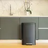 A dark gray pedal trash can is placed in front of gray kitchen cabinets and a marble-patterned backsplash with a faucet and a potted plant. Podgląd zdjęcia produktu