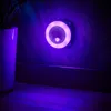 A night light with a motion sensor is plugged into a wall socket, casting a purple glow on the wall and floor. A plant in a white pot is visible on the left. Produktbild-Vorschau