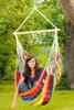 A woman is sitting in a colorful hammock chair hanging from a tree in a garden. Anteprima dell’immagine prodotto