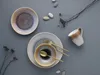 A flat lay of ceramic tableware and cutlery on a gray background, with decorative stones and dried plants. Náhľad obrázku produktu