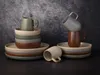A still life arrangement of ceramic plates and mugs in various earthy tones, set against a textured dark gray background. Produktbild-Vorschau