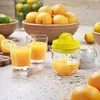 Two glasses of orange juice sit on a counter next to a manual juicer filled with orange juice. A bowl of oranges is in the background. Produktbild-Vorschau