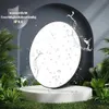 A round LED ceiling light with water splashes on it, displayed on a white podium surrounded by greenery. Podgląd zdjęcia produktu