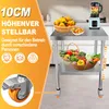 A stainless steel work table with two shelves, shown in a kitchen setting with fruits and vegetables. A blender is on the top shelf. The image highlights adjustable height and wheels. Produktbild-Vorschau