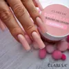 A close-up shot of a hand with long, light pink acrylic nails, next to a pink jar of Claresa builder gel. Small pink pom-poms are scattered around the jar. Produktbild-Vorschau