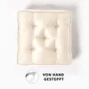 A square cushion with tufted details and a handle on top, presented on a white background with a graphic and text indicating it is hand-stitched. Náhled obrázku produktu