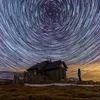 A person stands in front of a small wooden building under a sky filled with star trails. Náhľad obrázku produktu