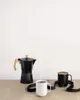 A black moka pot and two Ember mugs (one black, one white) are arranged on a wooden surface against a white background. Náhled obrázku produktu