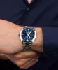 A man's wrist wearing a silver watch with a blue face, with his other hand resting on top. Produktbild-Vorschau
