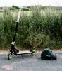 A scooter and helmet are placed on a paved surface next to tall grass. Produktbild-Vorschau