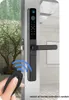 A hand holding a remote control is shown in the foreground, with a smart lock on a door in the midground. The background shows a blurred outdoor scene with trees and foliage. Náhled obrázku produktu