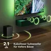 A soundbar and subwoofer are displayed in a modern living room setting, with a TV in the background and green ambient lighting. Voir les images du produit