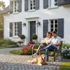 A couple and their dog relax on a garden bench in front of a house. Produktbild-Vorschau