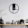 A modern, minimalist wall clock is displayed on a white wall above a wooden shelf with various decorative items and books. Podgląd zdjęcia produktu