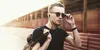 A young man wearing sunglasses and headphones, holding a backpack, walks outdoors. Anteprima dell’immagine prodotto