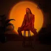 A woman in a red suit sits on a stool in front of a large, glowing orange circle projected onto a wall, with a plant to the left and a couch to the right. Produktbild-Vorschau