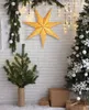 A festive Christmas scene with a large gold star decoration hanging on a white brick wall, flanked by two Christmas trees and gift boxes. Podgląd zdjęcia produktu