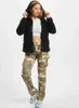 A woman is posing in a studio setting wearing a black hooded jacket and camouflage pants. Produktbild-Vorschau