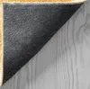 The underside of a rug is shown, folded over to reveal the textured backing and the edge of the rug. The rug is placed on a light-colored wooden surface. Produktbild-Vorschau