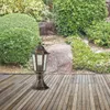 A garden lamp stands on a wooden deck in front of a lush garden. Produktbild-Vorschau