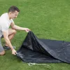 A man is kneeling on a grassy lawn, holding up a black tarp. Produktbild-Vorschau