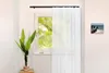 A window with white sheer curtains and a black curtain rod, with a plant in a vase on the left and a painting on the right wall. Anteprima dell’immagine prodotto