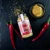 A bottle of chili mustard is displayed with red chili peppers and mustard seeds scattered around it on a dark surface. Produktbild-Vorschau