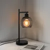 A black metal desk lamp with a cage-like shade and a visible Edison-style bulb sits on a white surface next to an open book. Podgląd zdjęcia produktu