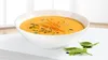 A bowl of carrot soup garnished with herbs and spices, placed on a white surface with a window in the background. Produktbild-Vorschau