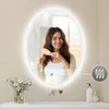 A woman is brushing her hair in front of an illuminated oval mirror in a bathroom setting. Náhled obrázku produktu