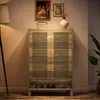 A wooden shoe cabinet with slatted doors is shown in a room setting, with a lamp on the left and a plant on the right. There are framed pictures and a vase on top of the cabinet. Produktbild-Vorschau