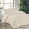A neatly made bed with cream-colored bedding is shown in a room with a large window and a plant. Produktbild-Vorschau