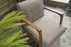 A close-up, slightly angled view of a cream-colored armchair with wooden armrests and frame, placed on a rug. A large green plant is in the foreground, partially obscuring the chair. Produktbild-Vorschau