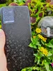 A hand holding a smartphone with a black honeycomb pattern on the back, surrounded by green plants and yellow flowers. Produktbild-Vorschau