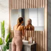 A woman is applying skincare in a modern bathroom with a wooden accent wall and a large illuminated mirror. Produktbild-Vorschau