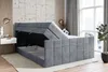 A modern bedroom with a gray upholstered bed frame, partially open to reveal storage space underneath. The bed is set against a textured wall with vertical wooden slats and a large window with curtains in the background. Produktbild-Vorschau