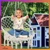A young girl sits in a white macrame hanging chair, giving a thumbs up, with a blue and red playhouse in the background. Voir les images du produit