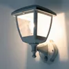 A white outdoor wall lamp with a motion sensor is mounted on a wall. Produktbild-Vorschau